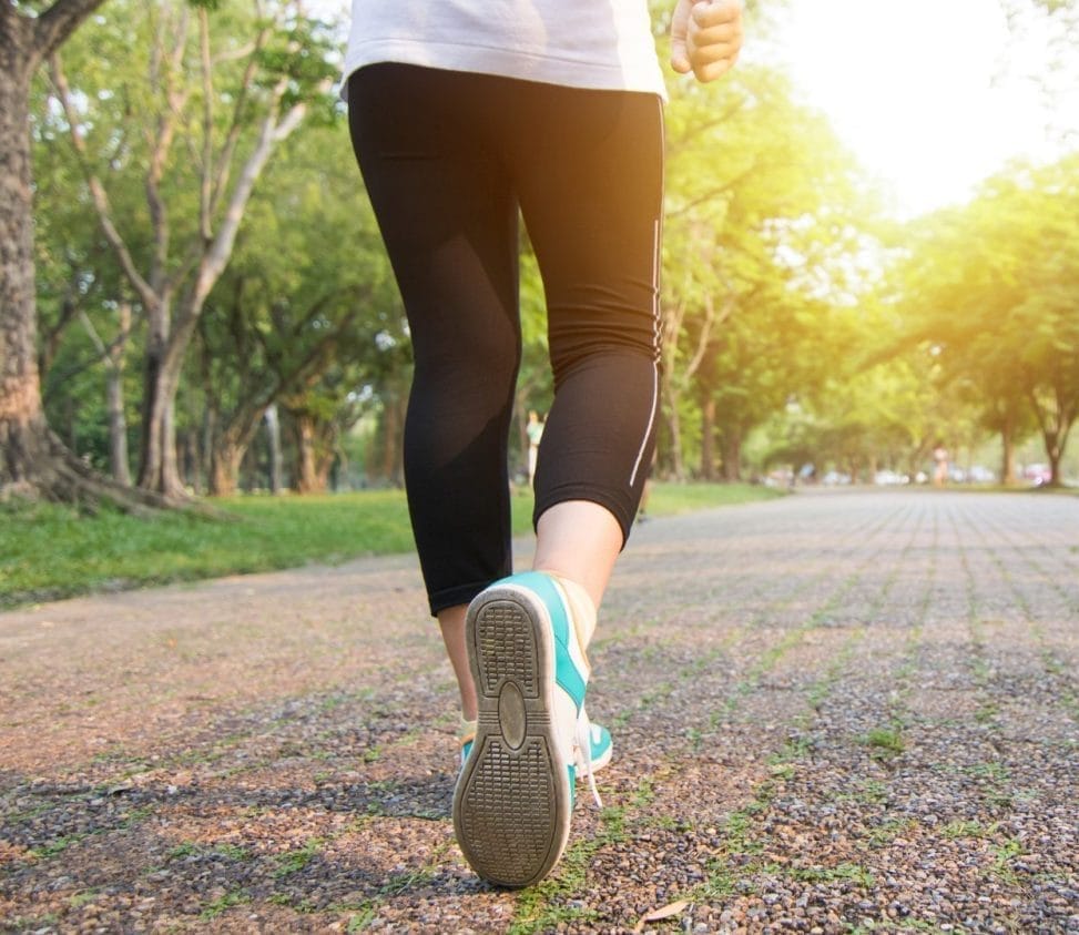 A person wearing black leggings and teal sneakers walks along a tree-lined path in a sunlit park, with green trees and sunlight visible in the background.