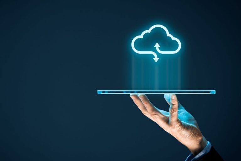 A hand holds a transparent tablet with a glowing cloud computing icon above it, symbolizing cloud storage and data transfer against a dark blue background.