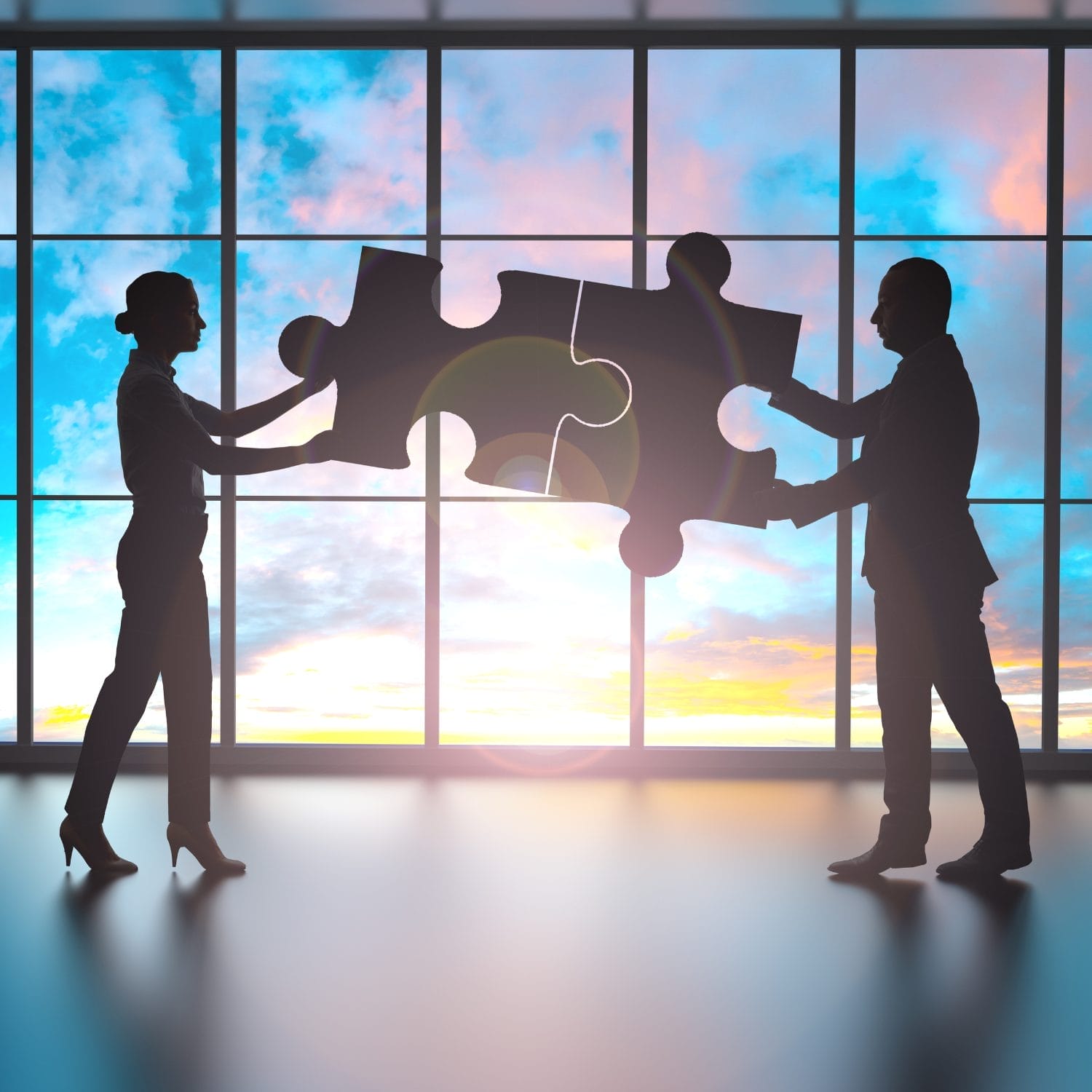 Two businesspeople, silhouetted against a large window with a colorful sunset sky, hold and connect two giant puzzle pieces facing each other.