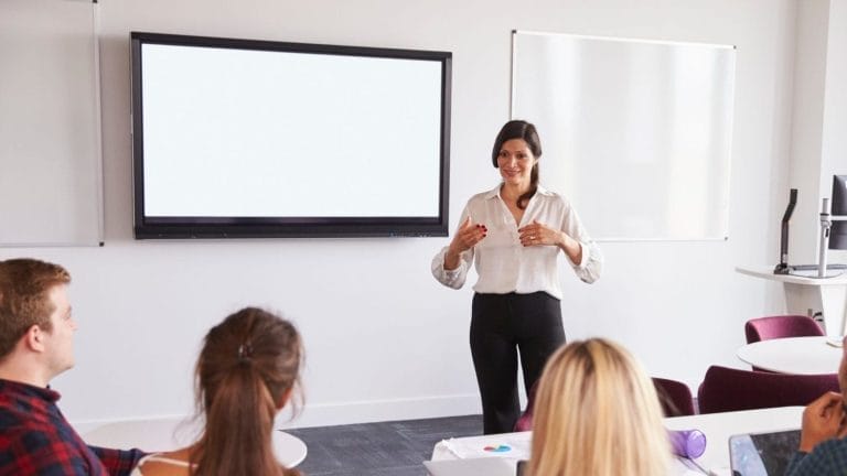 A woman stands in front of a large screen and whiteboard, speaking to a group of seated adults in a classroom or meeting room setting.