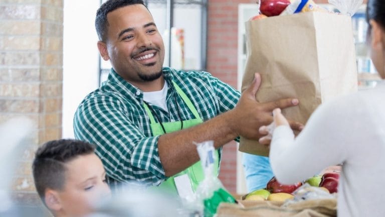 A smiling grocery store worker in a green apron hands a paper bag full of groceries to a customer. A child stands nearby at the checkout counter.