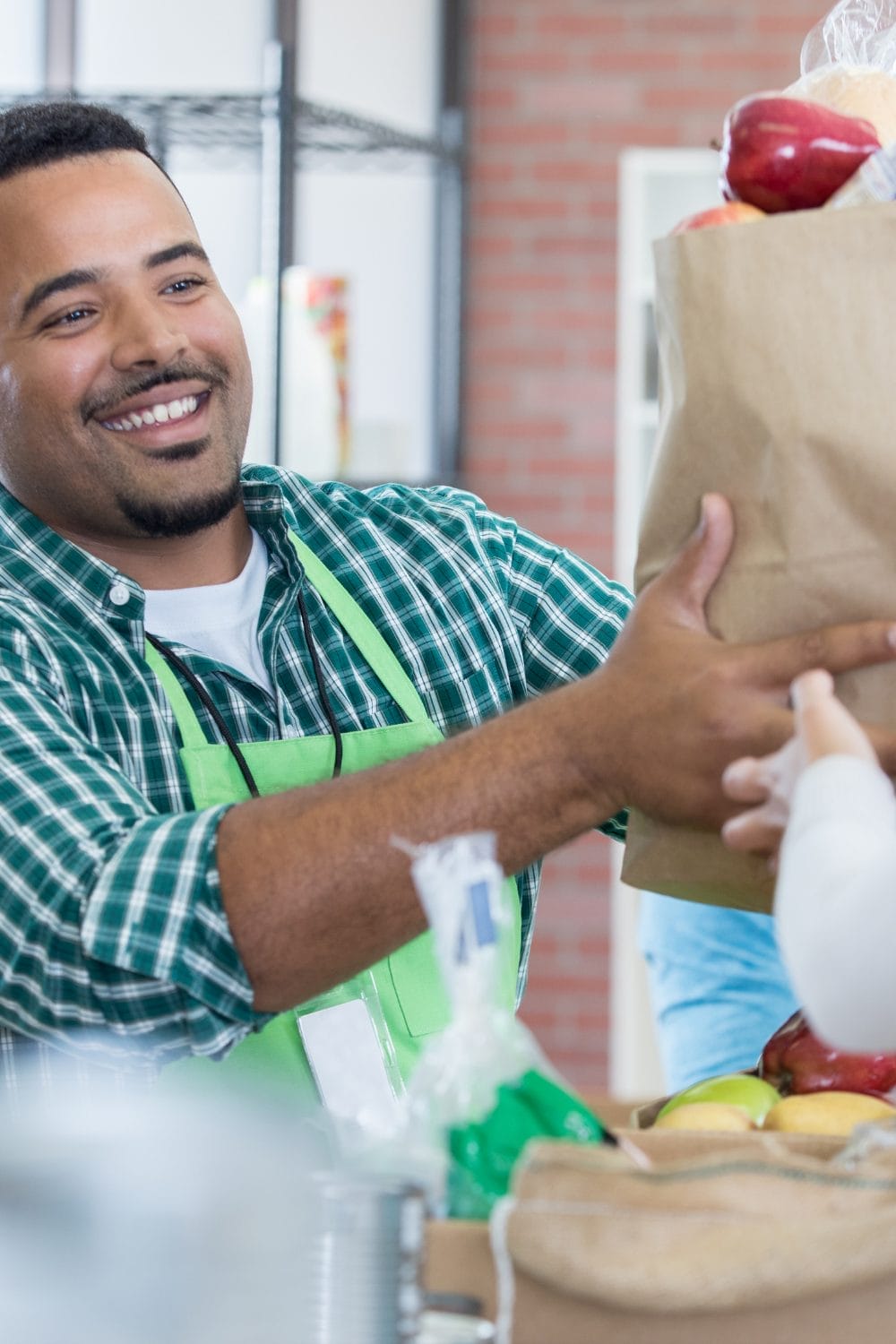 A smiling man in a green plaid shirt and apron hands a paper grocery bag filled with apples to someone across a counter in a store or market.