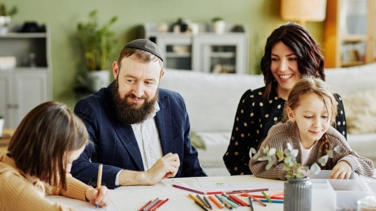 A bearded man wearing a kippah and a woman with dark hair smile while sitting at a table with two young girls who are drawing with colored pencils in a cozy, sunlit living room.