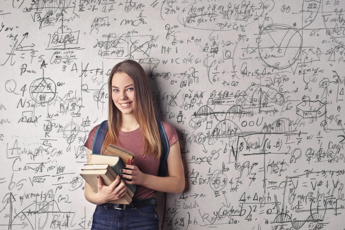 A young woman holding a stack of books stands in front of a whiteboard filled with complex mathematical equations and diagrams, smiling confidently—an inspiring image of higher education change management in action.