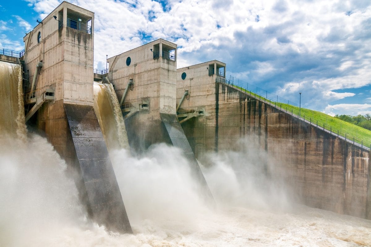 A concrete hydroelectric dam releases water through its spillways, creating mist and turbulent flow below, with blue sky and clouds overhead—much like how Nonprofit CRM costs explained helps clarify the flow of expenses for organizations.