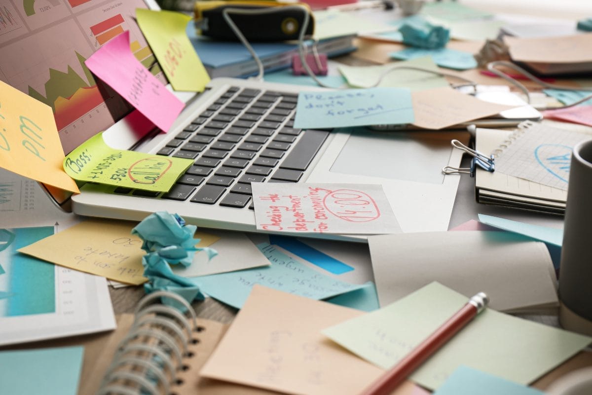 A cluttered desk with a laptop surrounded by colorful sticky notes, papers, notebooks, and office supplies, creating a chaotic and busy workspace.