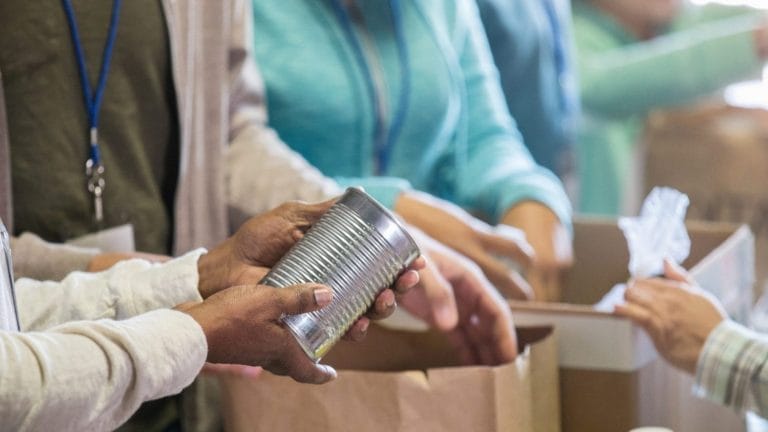 Close-up of people volunteering at a food bank, with one person holding a tin can and others packing items into paper bags. Only their hands and torsos are visible.