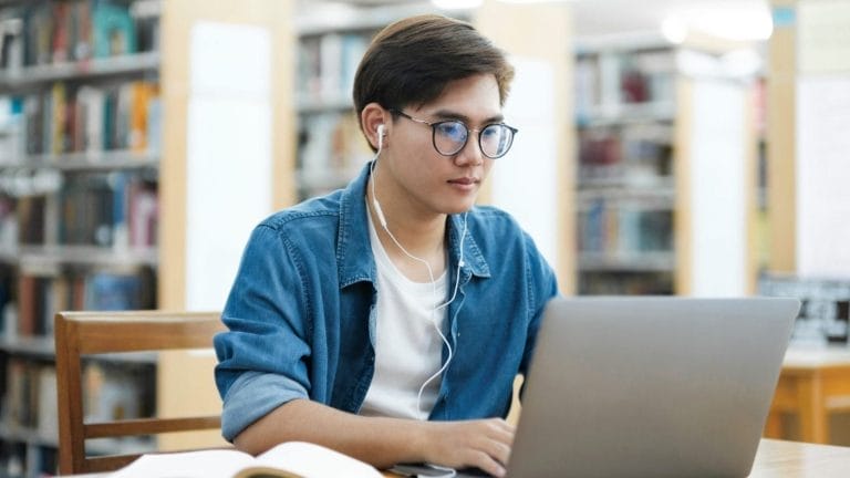 A young person wearing glasses and earphones sits at a desk in a library, focused on working on a laptop, with an open book in the foreground and bookshelves blurred in the background.