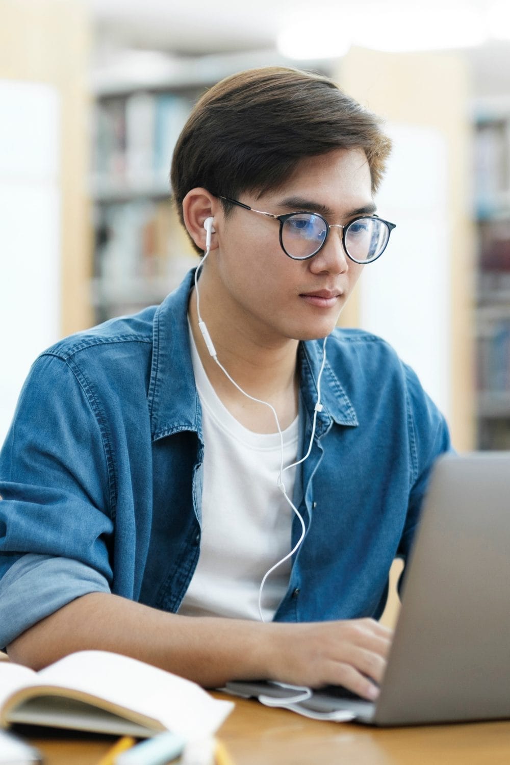 A young person wearing glasses and earphones is sitting at a desk in a library, focused on a laptop. An open book is visible in the foreground.