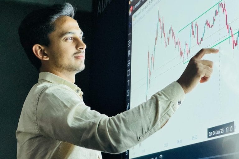A man in a beige shirt points at a screen displaying a financial chart with red and green candlesticks and trendlines, appearing to analyze or explain stock market data.