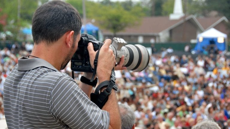 A man in a gray striped shirt uses a professional camera with a large lens to take photos at an outdoor event, with a large, blurred crowd in the background.