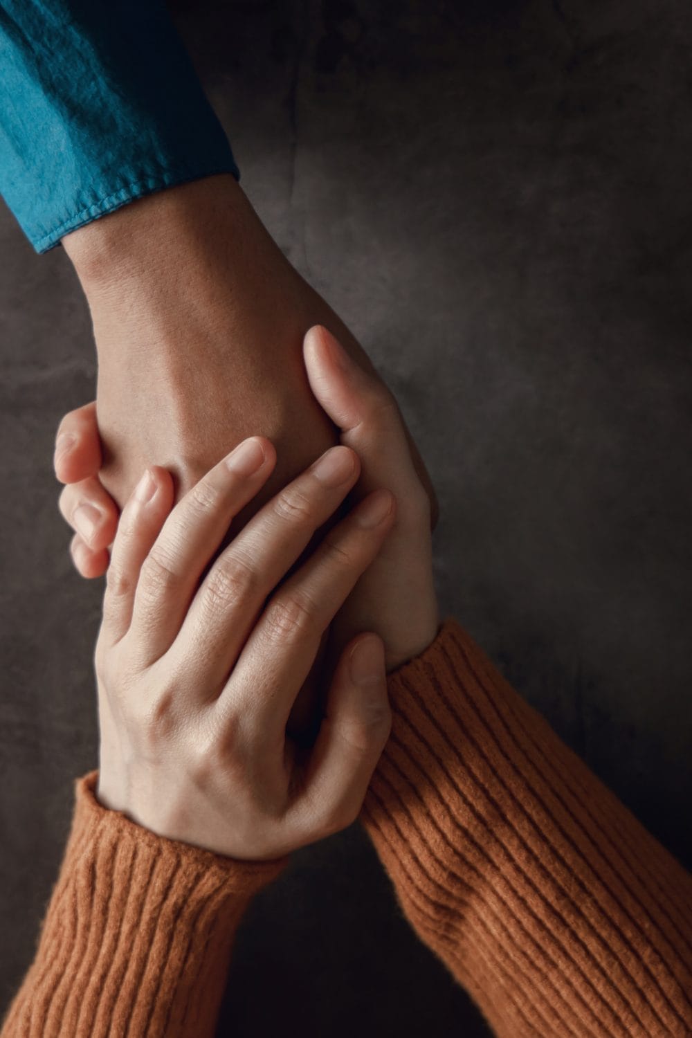 Two people holding hands; one hand in a blue sleeve is gently held by two hands in an orange-brown ribbed sweater against a dark background, conveying comfort and support.