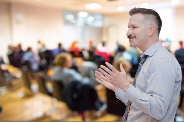 Jett Scott Winders in a dress shirt and tie speaks to an audience in a blurred conference room, gesturing with his hands while people sit and listen in the background.