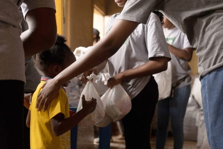 A group of people distribute plastic bags of supplies to a young child wearing a yellow shirt inside a room. One adult places a comforting hand on the childs shoulder.