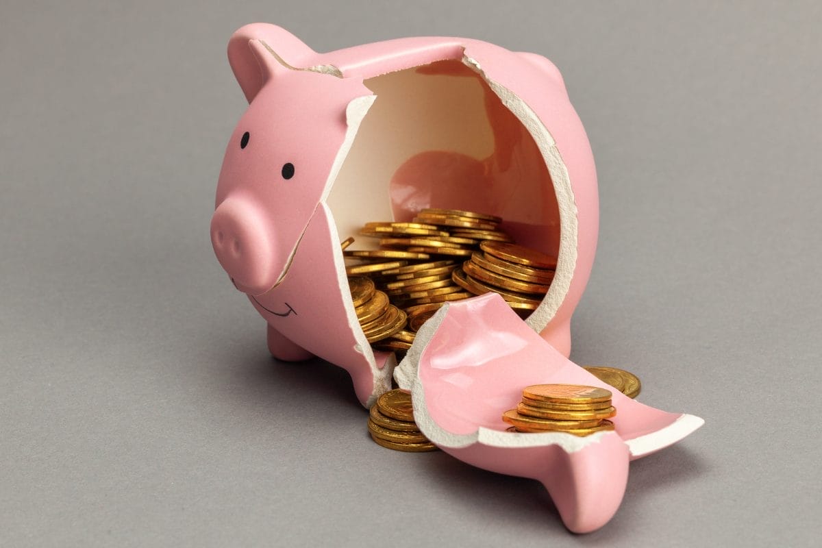 A broken pink piggy bank with gold coins spilling out onto a gray surface. The piggy bank is cracked open, and coins are visible both inside and outside the broken pieces.