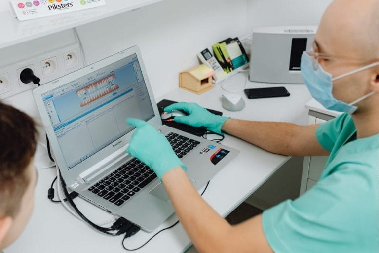 A person wearing gloves and a face mask points at a dental X-ray displayed on a laptop screen while discussing best practices for healthcare IT leaders during hospital mergers in a clinical office setting.