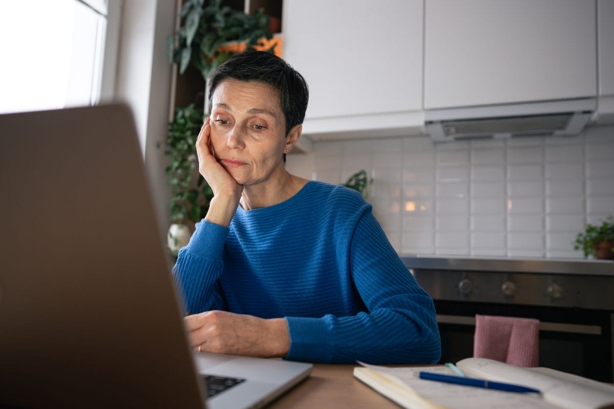 A woman in a blue sweater sits at a kitchen table, looking thoughtfully at a laptop screen with her chin resting on her hand. An open notebook and pen are on the table beside her.