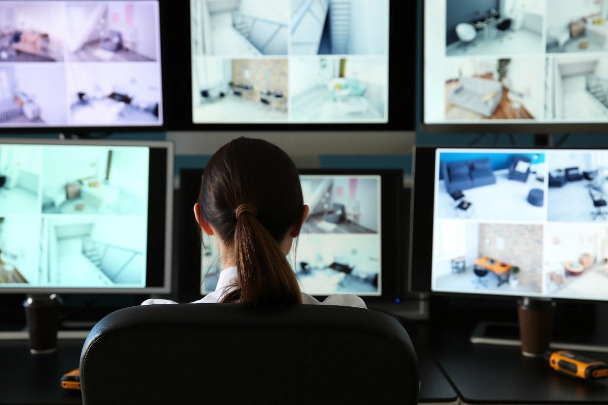 A person with brown hair in a ponytail sits at a desk, monitoring multiple security camera feeds showing various rooms on several screens in a control room.