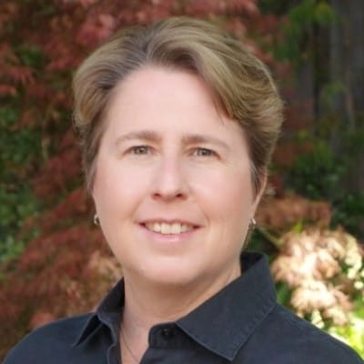 Joanne Jacobs Humane Society Silicon Valley A person with short, light brown hair smiles outdoors, wearing a dark collared shirt. Soft greenery and reddish foliage are blurred in the background.