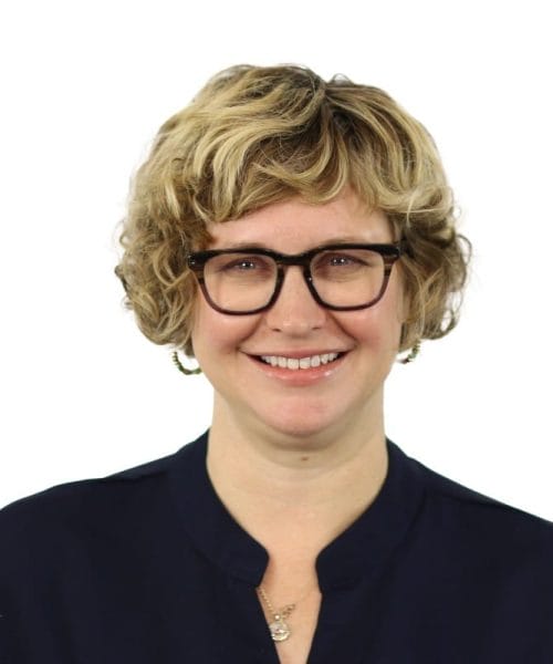 Noel Radley 2025 A smiling person with short, curly blonde hair and black glasses, wearing a dark navy top and a necklace, posed against a plain white background.