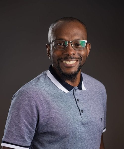 Okiemute Numa 2025 Heller Headshots – Delaney Rain Photography-29 A smiling man with glasses, a goatee, and short hair wears a light blue polo shirt with dark trim, standing against a plain dark background.