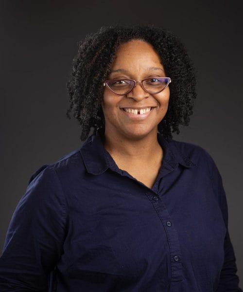 Shawneda Crout 2025 Heller Headshots – Delaney Rain Photography-41 Heller Consulting President Jeffrey Appell, with curly hair and glasses, smiles at the camera in a dark navy button-up shirt, standing against a plain dark background.