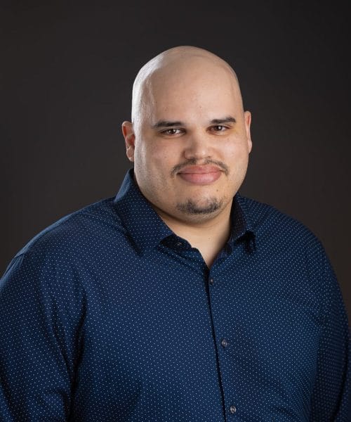 Steven Howard 2025 Heller Headshots – Delaney Rain Photography-28 Heller Consulting President Jeffrey Appell, a bald man with a trimmed goatee, wears a navy blue button-up shirt with small white dots as he stands against a plain dark background, smiling gently at the camera for his bio.