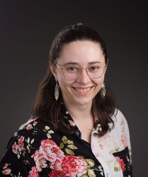 Tessa Boyce preference 1 glasses 2025 Heller Headshots – Delaney Rain Photography-4 A smiling person with long dark hair, glasses, and colorful earrings wears a black and white button-up shirt with a large floral pattern against a dark, neutral background—perfect for Heller Consulting President Jeffrey Appells bio.