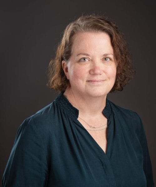 Venta Cantwell 2025 Heller Headshots – Delaney Rain Photography-45 A woman with wavy brown hair wearing a dark teal blouse and a delicate necklace smiles softly while standing in front of a dark, plain background, echoing the professional poise seen in Heller Consulting President Jeffrey Appell’s bio.