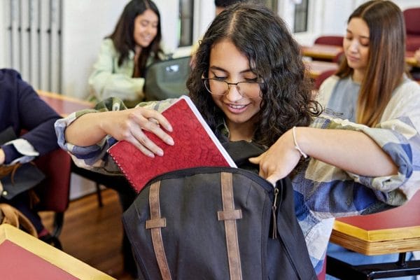student gordon college A student with curly hair and glasses smiles while placing a red notebook into her backpack in a classroom, with other students sitting at desks in the background.